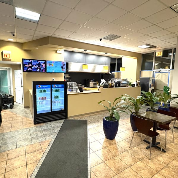 Interior view of a cafe with a counter, menu screens, Pepsi coolers, and seating area.