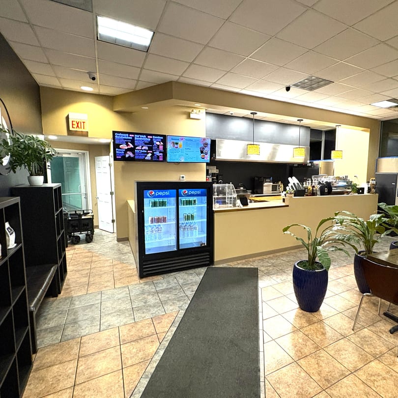 Interior view of a cafe with a counter, menu screens, Pepsi refrigerators, and potted plants.