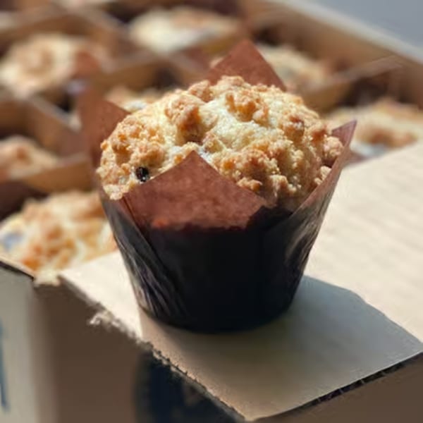 A blueberry muffin with a crumb topping in a brown paper liner, sitting on a cardboard box.