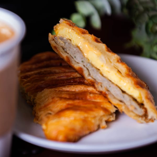 A golden-brown pastry, cut in half, revealing layers of egg and sausage, next to a coffee cup.