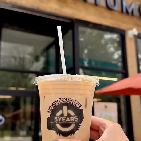 Hand holding an iced coffee from Momentum Coffee, with the storefront visible in the background.