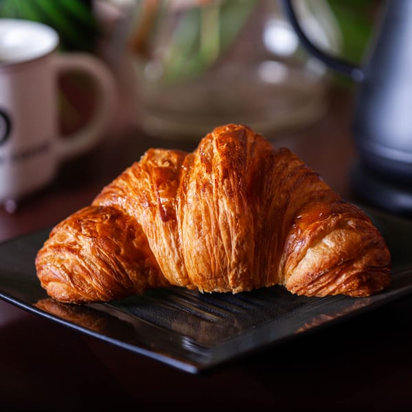 Golden-brown croissant on a dark plate, with a coffee mug and blurred background at Momentum Coffee.