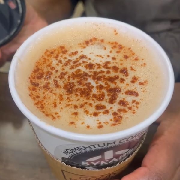 A Momentum Coffee latte with foamed milk and cinnamon sprinkles, held by hands.