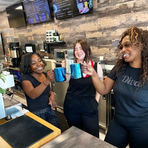 Three smiling Momentum Coffee baristas toast with blue and silver mugs behind the counter.